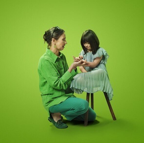 Two people wearing green. A little girl is seated on a stool while another woman kneels and holds her hand against a green background.