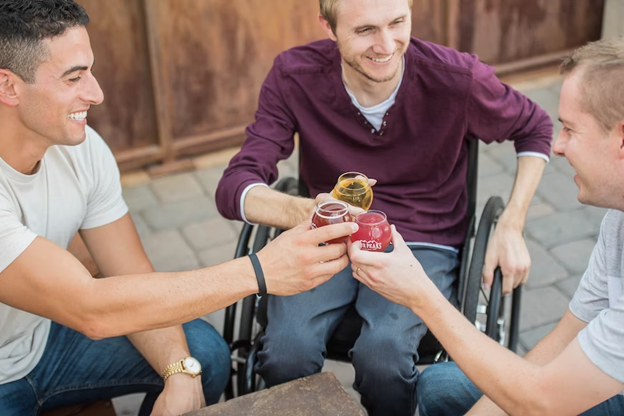 Two men and a man in a wheelchair are raising their drinks in celebration.