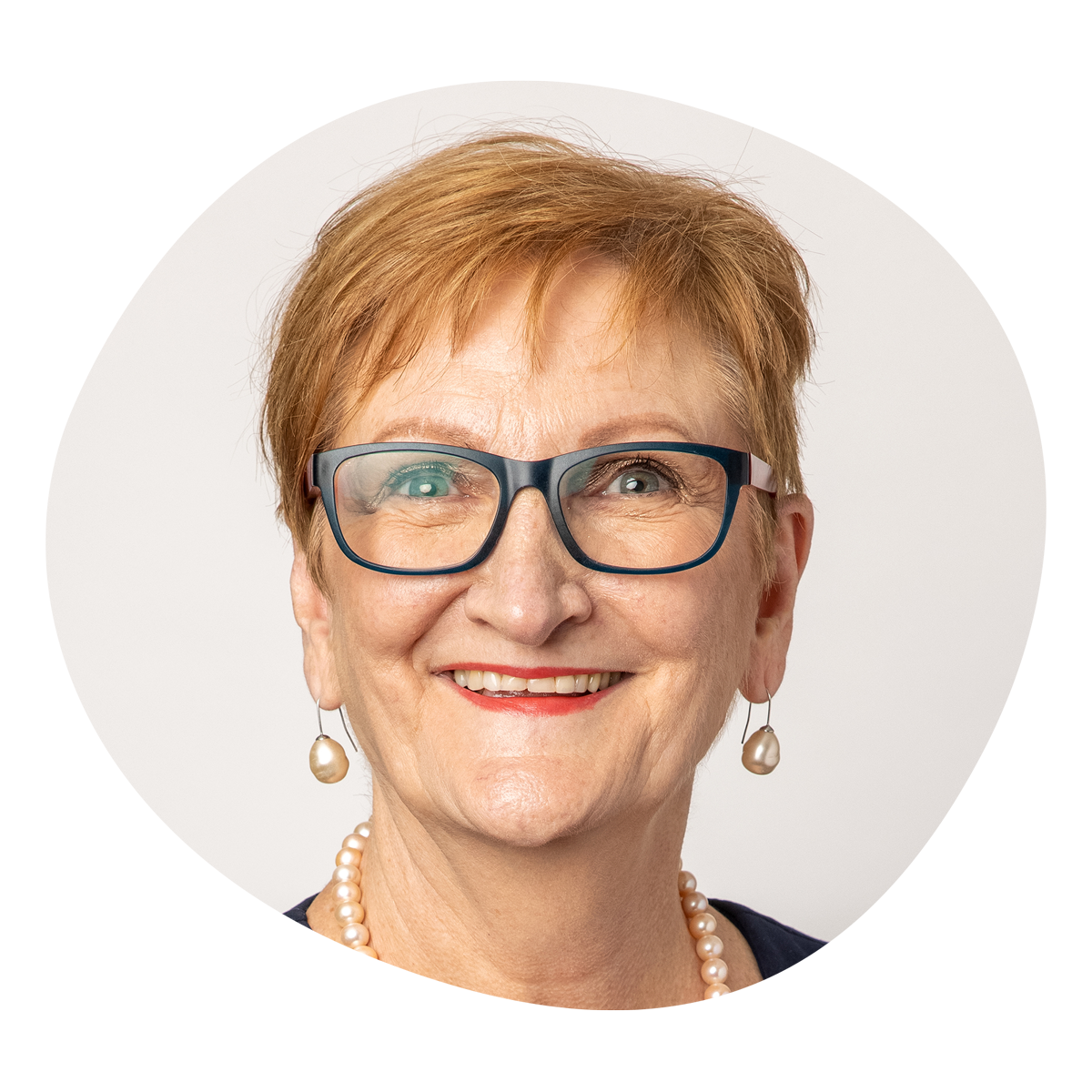 Elderly person smiling warmly, wearing large blue glasses, pearl earrings, and necklace. Short, light brown hair. Neutral background, cheerful tone.