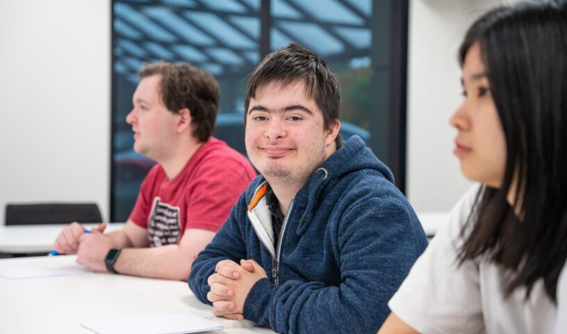 Young man sitting at a desk smiling to camera.